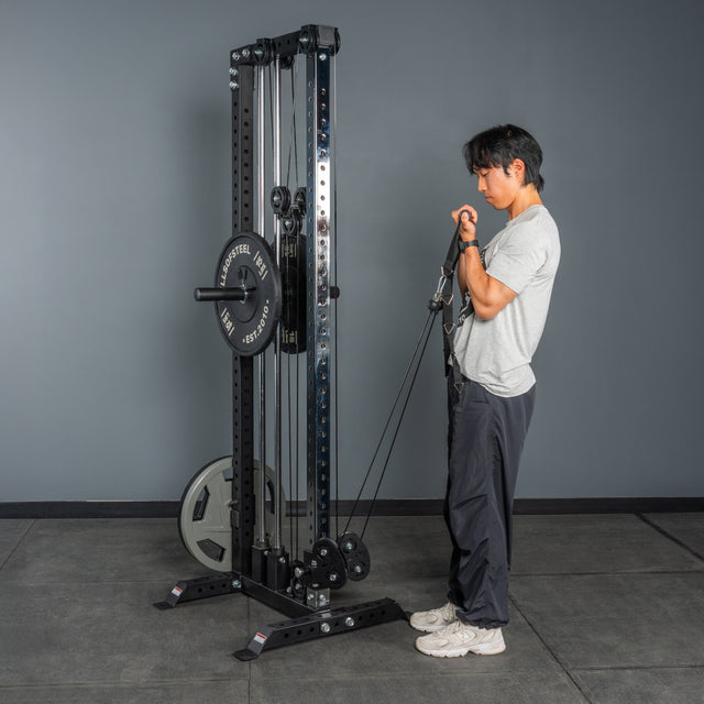 A person in a gray t-shirt and dark pants performs bicep curls on the Bells of Steel Cable Tower in a gym with gray walls and a black floor.