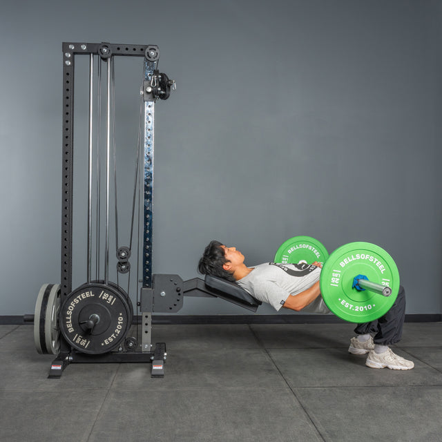 Wearing athletic gear, a person does a hip thrust with a barbell loaded with green plates, using a bench and the Bells of Steel Cable Tower's weight stack for support in the gym.