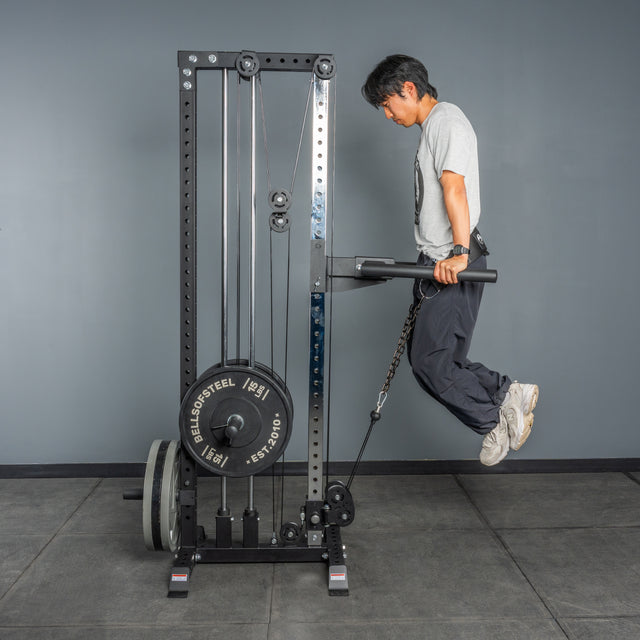 A person performs dips on the Bells of Steel Cable Tower, using its adjustable weight stack, gripping the handles with arms extended and legs bent backward in a gym.