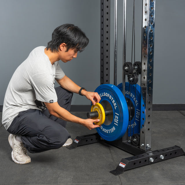 Wearing athletic gear, a person kneels to load a yellow weight plate onto the Bells of Steel Cable Tower, which already holds blue plates, preparing for cable machine exercises in the gym.