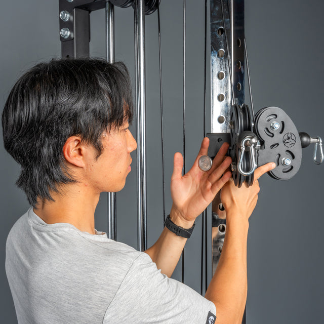 Wearing a gray shirt, a person adjusts the Bells of Steel Cable Tower with a metal attachment and coin near the weight stack, against a gray backdrop—getting ready for effective cable workouts.