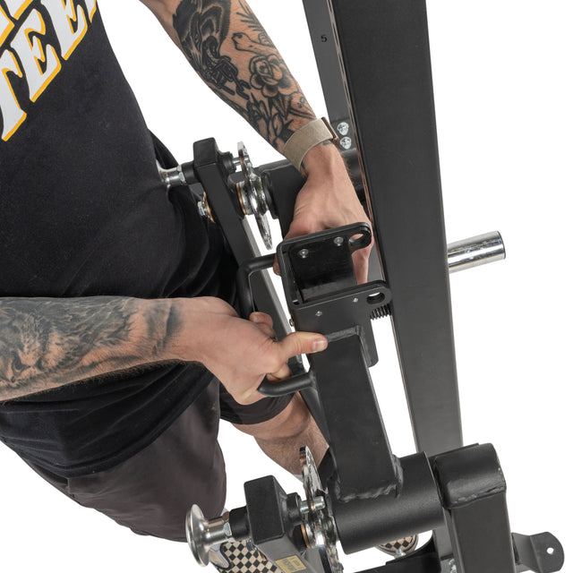 A tattooed individual grips the Bells of Steel Shoulder Boulder/Chest Fly Attachment, preparing to use it. The overhead image captures part of the equipment and their torso.