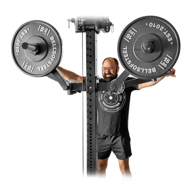 A man in a black t-shirt and shorts smiles behind the Bells of Steel Shoulder Boulder/Chest Fly Attachment, ready for a chest fly workout, with large branded weight plates beside the power rack.