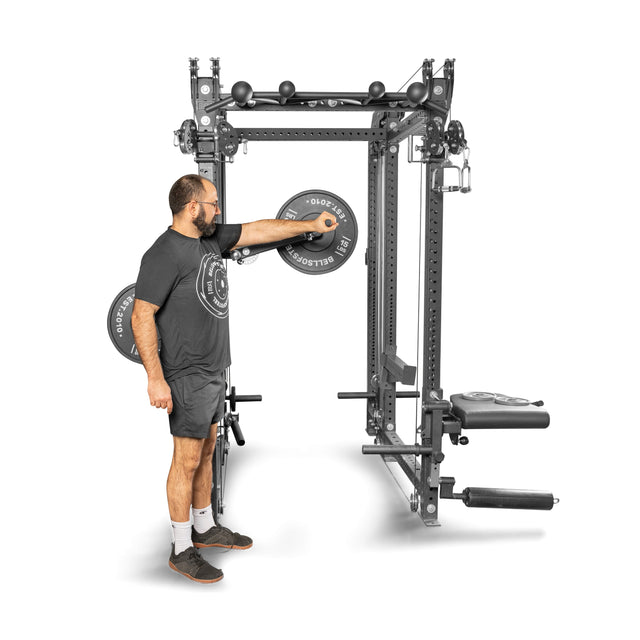 A man in a black t-shirt, shorts, and mask adjusts weights on a Bells of Steel gym machine featuring the Shoulder Boulder/Chest Fly Attachment; a bench is beside him against a plain white background.