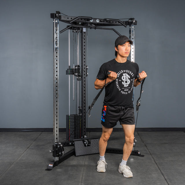 A person in a black shirt, black shorts, and cap uses the Bells of Steel All-in-One Trainer for resistance exercises in a gym with gray walls and a black floor.