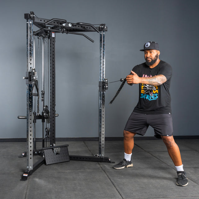 A man in a black T-shirt, shorts, and cap works out at the gym, using the Bells of Steel All-in-One Trainer to pull the cable handle across his body with both hands while standing.