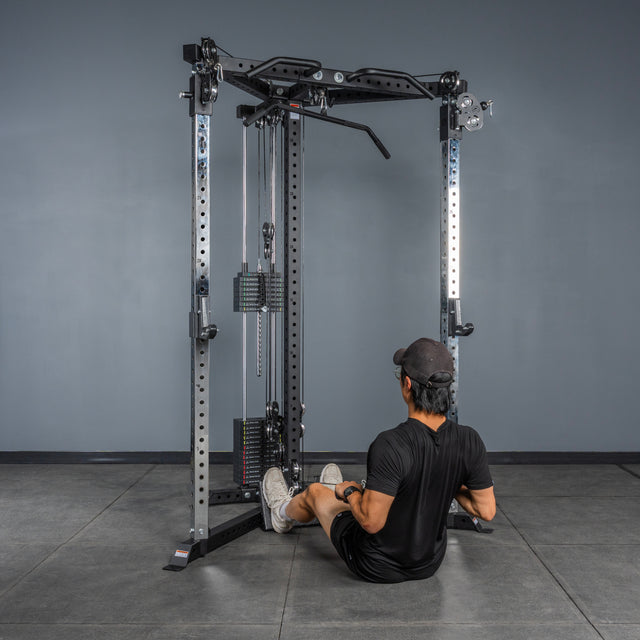 Dressed in black workout gear and a cap, a person sits on the tile floor of their home gym with gray walls, preparing for a seated row using the Bells of Steel All-in-One Trainer.
