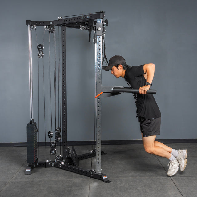 A man in athletic wear and a cap uses the Bells of Steel All-in-One Trainer at the gym, performing a tricep pushdown with both hands on a straight bar while leaning forward.