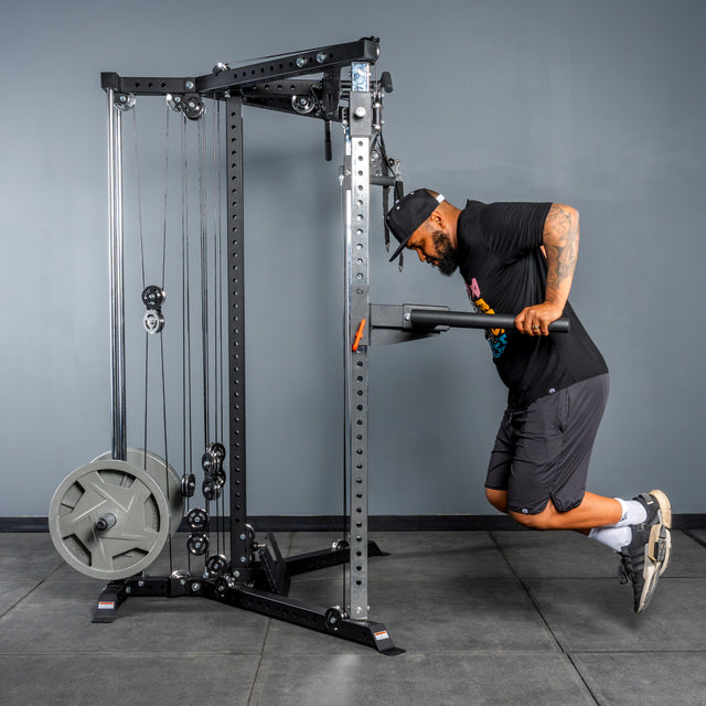 A man in a black hat, t-shirt, and shorts does triceps dips on the Bells of Steel All-in-One Trainer in a gym with gray walls and a concrete floor.