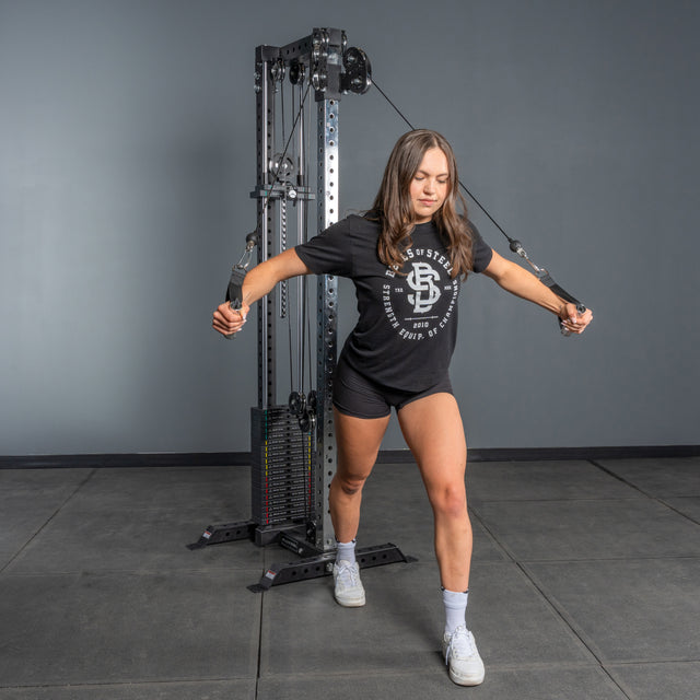 A woman in a black t-shirt and shorts uses the Bells of Steel Cable Tower at the gym, performing a cable crossover with extended arms and one leg forward in a lunge position.