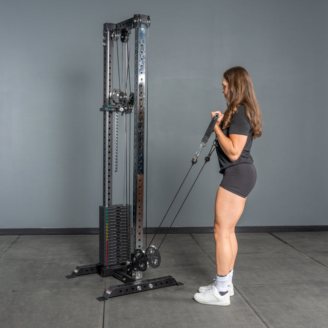 A woman in athletic wear stands before the Bells of Steel Cable Tower, gripping the handles and getting ready to perform cable machine exercises in a gym.