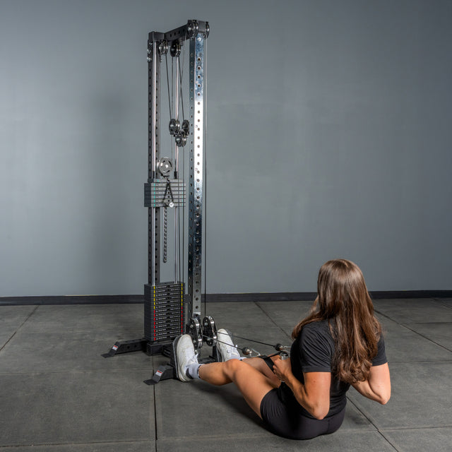 A woman with long brown hair sits on a gym floor facing the Bells of Steel Cable Tower, holding its handles and preparing for cable exercises. The room features gray walls and a smooth, tiled floor.