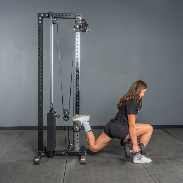 A woman does a Bulgarian split squat with dumbbells, elevating one foot on the Bells of Steel Cable Tower, demonstrating an effective cable machine exercise in the gym.