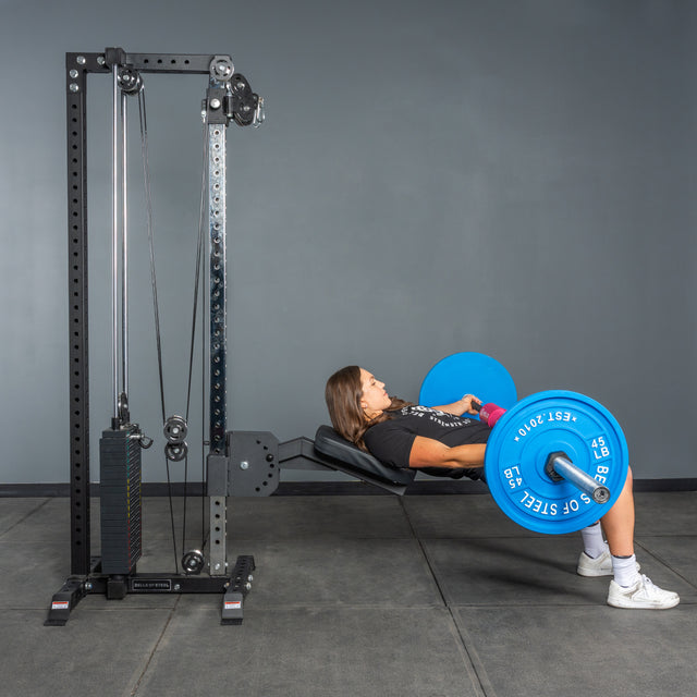 A woman performs a barbell hip thrust on a bench in the gym using a blue weight plate, with the Bells of Steel Cable Tower and its weight stack visible nearby.