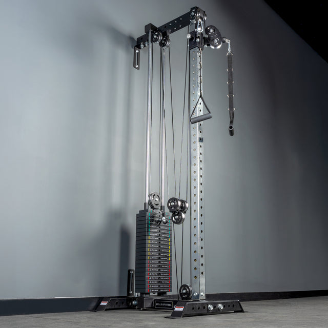The Bells of Steel Cable Tower is positioned against a gray wall on a gray floor. Featuring metal weight stacks, pulleys, rope handle, and attachments, it’s ideal for various cable machine exercises.