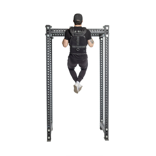 A man in a black cap and workout clothes performs a pull-up using a Bells of Steel Weighted Vest on a metal bar, showcasing his strength from behind with legs slightly bent, enhanced by the vest's adjustable training plates.
