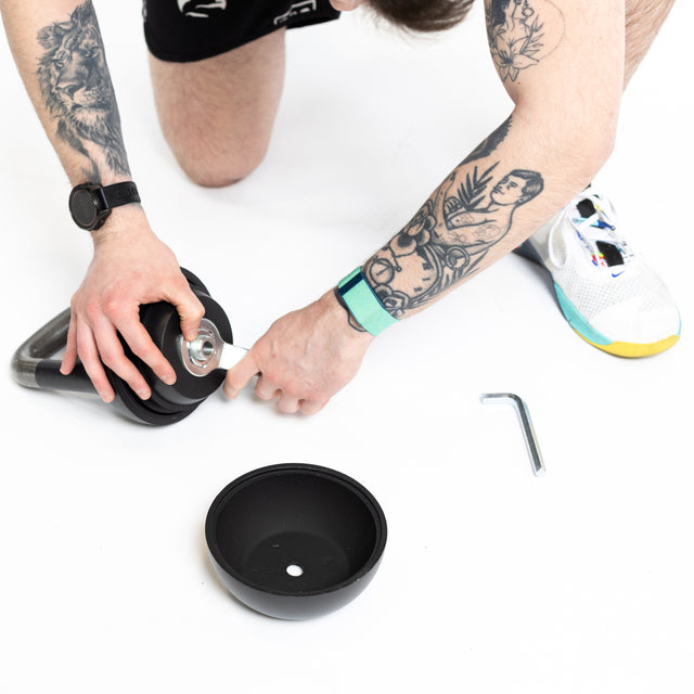 A tattooed person with a smartwatch assembles a Bells of Steel 6-12KG Adjustable Kettlebell on a white surface, using a screwdriver and Allen wrench, with an extra weight plate nearby.