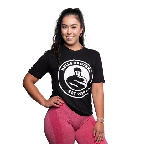 Woman with long dark hair wearing a black "Bells of Steel" T-shirt and pink leggings, standing with one hand on her hip and smiling against a plain white background.
