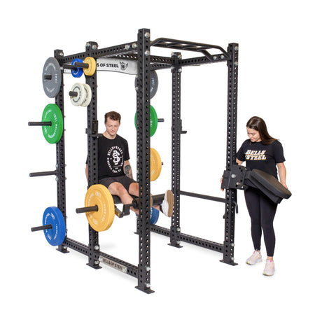 A man uses a squat rack with weighted plates for a leg exercise while a woman stands nearby holding a black padded attachment. Both wear black "Bells of Steel" shirts.