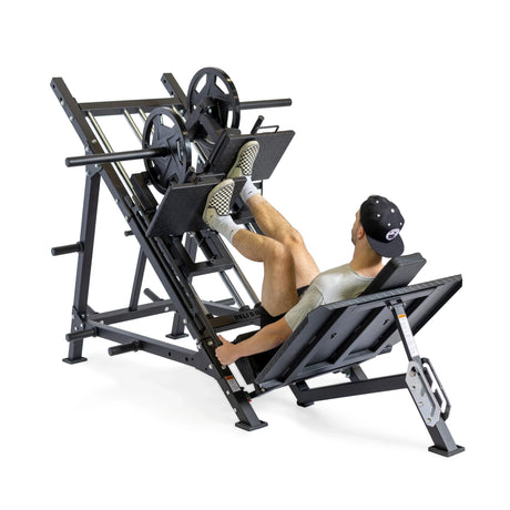 A man wearing a gray shirt, black shorts, and a cap uses a leg press machine loaded with weights, pushing the platform upward with his feet.