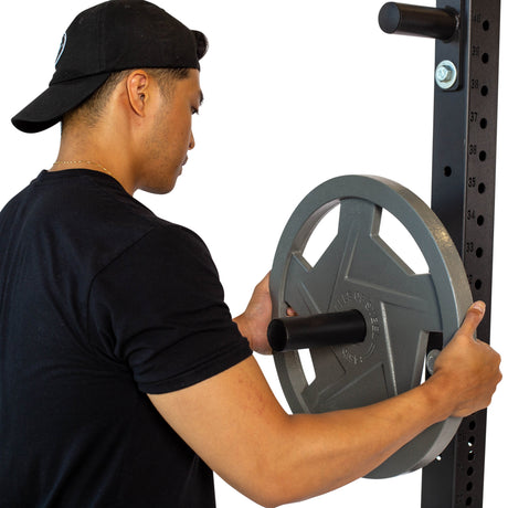 A person wearing a black shirt and cap places a large gray weight plate onto a barbell storage rack attached to a metal frame.