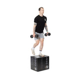 A person performs step-ups with dumbbells on a black Bells of Steel Foam Soft Plyo Box (16 x 18 x 20), wearing a black T-shirt, gray shorts, white socks, and sneakers against a plain white background.