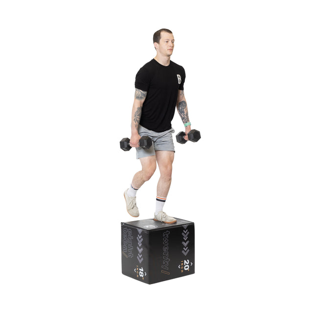 A person performs step-ups with dumbbells on a black Bells of Steel Foam Soft Plyo Box (16 x 18 x 20), wearing a black T-shirt, gray shorts, white socks, and sneakers against a plain white background.