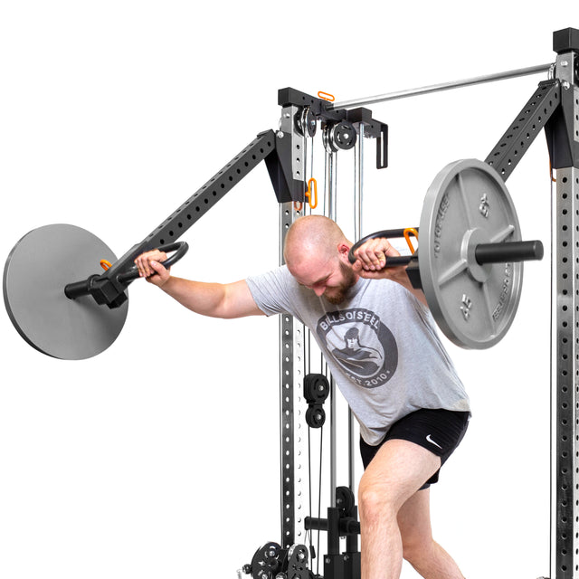 Wearing a Bells of Steel "Balls of Steel" tee and shorts, someone does a landmine squat with a barbell set on Cable Tower Squat Stands, gripping the bar with both hands and squatting with one knee bent.