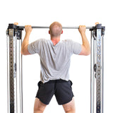 A man in a gray shirt and black shorts does a pull-up on the Bells of Steel Cable Tower Squat Stands, viewed from behind against a white background.