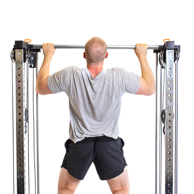 A man in a gray shirt and black shorts does a pull-up on the Bells of Steel Cable Tower Squat Stands, viewed from behind against a white background.