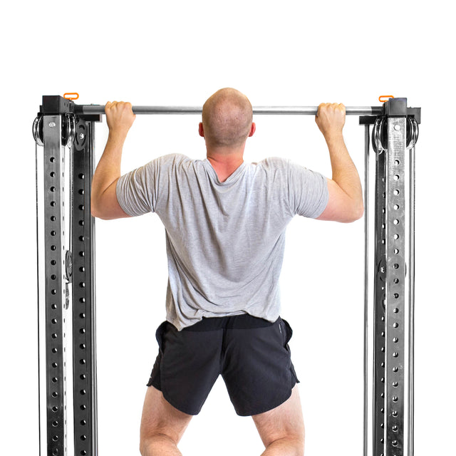A man in a gray t-shirt and black shorts does a pull-up on the Bells of Steel Cable Tower Squat Stands, seen from behind against a white background.
