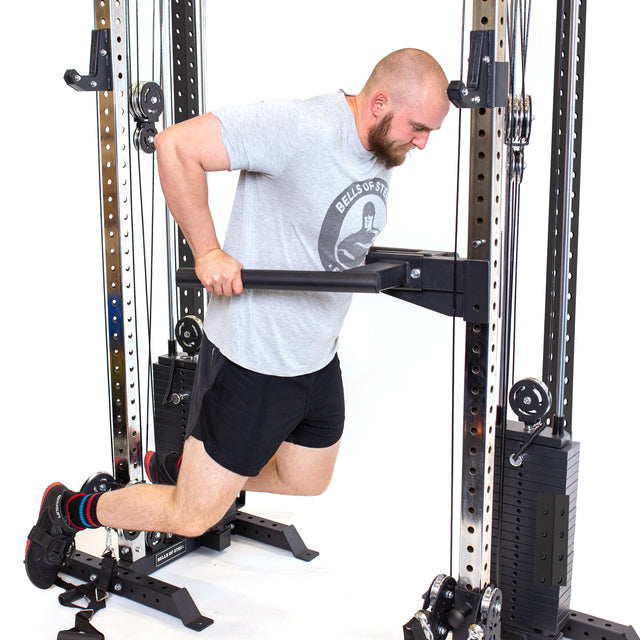 A person in gray t-shirt and black shorts does dip exercises with a dip bar on the Bells of Steel Cable Tower Squat Stands in a gym.
