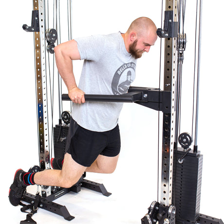 A man in a gray t-shirt and black shorts performs dips on Bells of Steel Cable Tower Squat Stands with a dip bar attachment, against a white background.