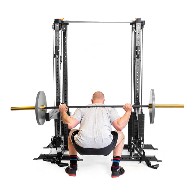 A person in a gray shirt and black shorts performs a barbell squat inside a power rack, with Bells of Steel Cable Tower Squat Stands positioned nearby and weight plates on both ends of the barbell, viewed from behind.