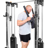 A man in a gray t-shirt and black shorts uses the Bells of Steel Cable Tower Squat Stands at the gym, holding both handles while facing the equipment.