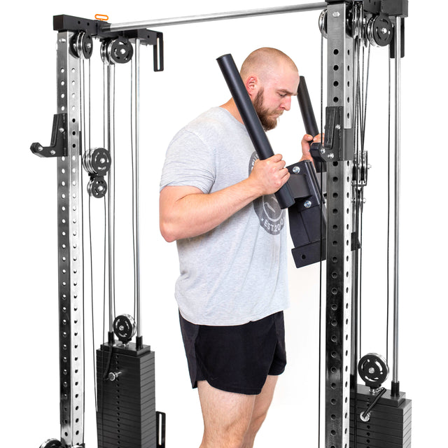 A man in a gray t-shirt and black shorts uses the Bells of Steel Cable Tower Squat Stands at the gym, holding both handles while facing the equipment.