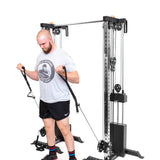 A man uses the Bells of Steel Cable Tower Squat Stands to perform bicep curls with resistance bands in a gym, wearing a gray T-shirt and black shorts.