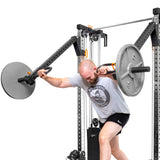 A man in a gray t-shirt and black shorts uses Bells of Steel Cable Tower Squat Stands to lift a barbell overhead while working out in a gym.