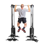 A man in a gray t-shirt, black shorts, and colorful socks performs a pull-up on the Bells of Steel Cable Tower Squat Stands, photographed from behind against a white background.
