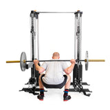 A person in a gray t-shirt and black shorts is performing a barbell squat from behind using Bells of Steel Cable Tower Squat Stands. The black squat stands hold the barbell, which has metal plates on each end.