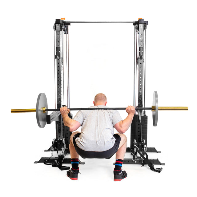 A person in a gray t-shirt and black shorts is performing a barbell squat from behind using Bells of Steel Cable Tower Squat Stands. The black squat stands hold the barbell, which has metal plates on each end.