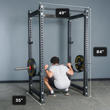 A person squats with a barbell inside a black Bells of Steel Manticore Four Post Power Rack - Prebuilt (3" x 3", 1" Holes), which measures 49 inches wide, 35 inches deep, and 84 inches tall. This sturdy power rack stands in a room with gray walls and a concrete floor.