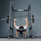 A person lies on a bench and grips a barbell with weights, preparing to bench press inside the Manticore Four Post Power Rack - Prebuilt (3" x 3", 1" Holes) by Bells of Steel in a gym setting.