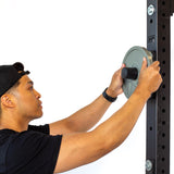A person in a black shirt and cap loads an Olympic-sized weight plate onto Bells of Steel Bolt-On Plate Pegs attached to a power rack, against a plain white background.