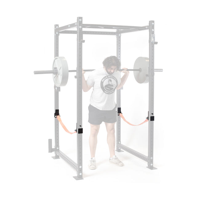 Wearing athletic clothing, a person squats with a barbell in a power rack featuring brightly colored Bells of Steel Safety Strap Connectors, which stand out vividly as both the person and equipment are partially faded.