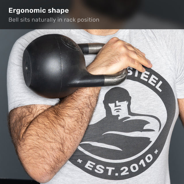A person holds a Bells of Steel 6-12KG Adjustable Kettlebell, competition-style and black in color, in the rack position against their chest. They wear a gray t-shirt featuring a graphic of a muscular figure and the text “STEEL EST. 2010.”.