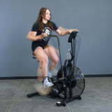 A woman rides the Bells of Steel Residential Air Bike with Wind Guard indoors. Her quick movements blur against a minimalist home gym backdrop with a gray wall and concrete floor.
