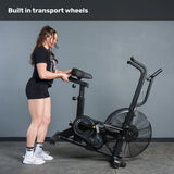 A woman in workout gear moves the Bells of Steel Residential Air Bike with Wind Guard using its built-in transport wheels. The text above reads, "Built in transport wheels"—ideal for any home gym.