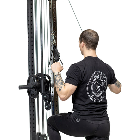 A tattooed person in a black "Strength Equipment" shirt uses Bells of Steel Adjustable Length Aluminum Handles on a cable machine for a lat pulldown, kneeling with a white background that highlights these premium gym accessories.