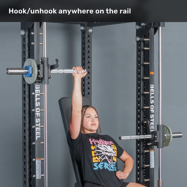 A woman seated at a Smith machine uses the Bells of Steel Anvil Handles to bench press a barbell with one arm raised. Text above reads, “Hook/unhook anywhere on the rail.” The machine is labeled “Bells of Steel.”.
